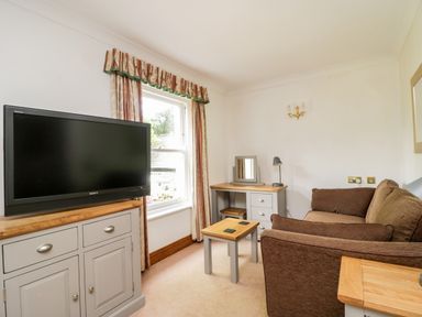 A small sitting room with a brown sofa a flat screen TV on a cabinet a window and a desk with a mirror and lamp at Thirlmere Suite in Thirlmere