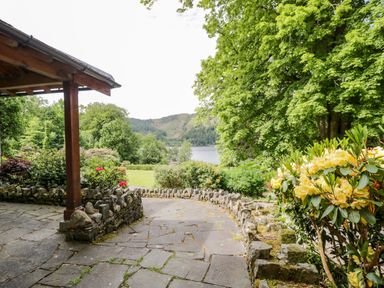 A stone patio with a wooden post and stone wall surrounded by green bushes and trees with a lake and hills in the background at Thirlmere Suite in Thirlmere