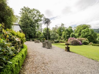 An outdoor gravel patio with wicker chairs surrounded by green bushes trees and a lawn with flowering shrubs at Thirlmere Suite in Thirlmere