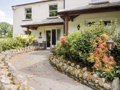 A stone walkway with rock borders leading to a white building entrance with two outdoor chairs and bushes on both sides at Thirlmere Suite in Thirlmere
