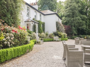 A white building with a porch surrounded by green hedges and flowering bushes with benches and outdoor wicker chairs on gravel at Thirlmere Suite in Thirlmere