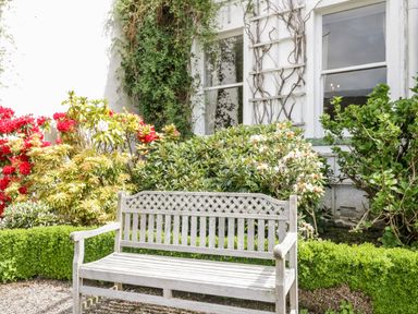 A wooden bench in a garden with shrubs and flowers near a white building with two windows at Thirlmere Suite in Thirlmere