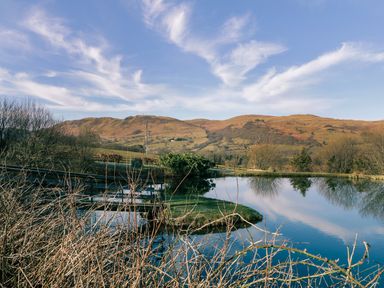 A lake with stone bridges and bare bushes in front of hills under a blue sky at Scales Cottage in Lorton near Cockermouth