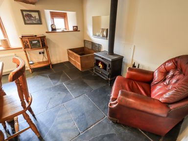 A room with a wood burning stove a red leather armchair wooden chairs and a wooden stand with a radio at Scales Cottage in Lorton near Cockermouth