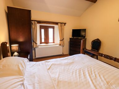 A bedroom with a wooden bed a nightstand with a lamp a window with curtains a dresser with a television and a chair at Scales Cottage in Lorton near Cockermouth