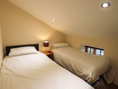 A bedroom with two single beds a small wooden nightstand with a lamp and remote control and a small window at Scales Cottage in Lorton near Cockermouth