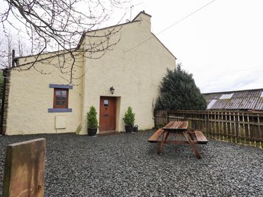 An outdoor area with a gravel ground a wooden picnic table a fenced boundary and a house wall with a wooden door and window at Scales Cottage in Lorton near Cockermouth