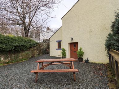 An outdoor area with a wooden picnic table in front of a cream house with a wooden door and stone wall at Scales Cottage in Lorton near Cockermouth