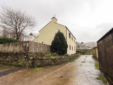 A paved path leading past a cream cottage with stone walls and a wooden fence at Scales Cottage Lorton near Cockermouth