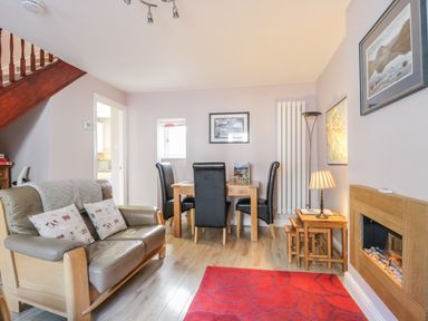 A living room with a leather loveseat, black dining chairs at a wooden table, a red rug, wall-mounted fireplace, and staircase at Swinside Cottage in Keswick