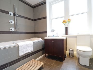 A bathroom with a bathtub a sink with a cabinet a toilet a trash can and a wooden stool at Keswick Loft in Keswick