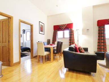 A dining area with a table and six chairs next to a living space with two black sofas and patterned curtains at Keswick Loft in Keswick