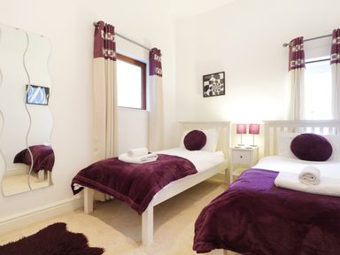 A bedroom with two single beds with purple blankets and pillows a small nightstand with two lamps between the beds and two windows with curtains at Keswick Loft in Keswick