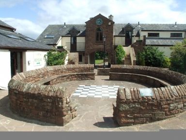 An outdoor circular stone seating area with a checkered floor and buildings with stone roofs in the background at Whitbarrow Holiday Village Troutbeck 5 Ullswater