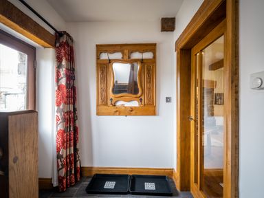A hallway with a wooden framed mirror on the wall two black trays on the floor a wooden door with glass and a window with red and gray floral curtains at Grayrigg Foot Stable in Grayrigg