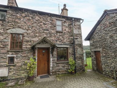 Front view of a stone cottage with a wooden door and windows with plants in pots on either side at Tanner Croft Cottage in Grasmere