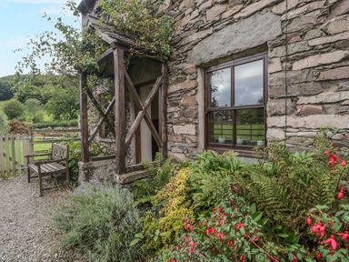 The exterior of a stone cottage with a wooden bench plants and a wooden framed window at Tanner Croft Cottage in Grasmere