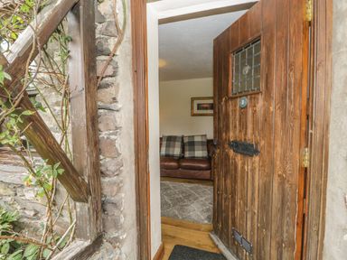 An open wooden front door showing a glimpse of a living room with a brown leather sofa and plaid cushions at Tanner Croft Cottage in Grasmere