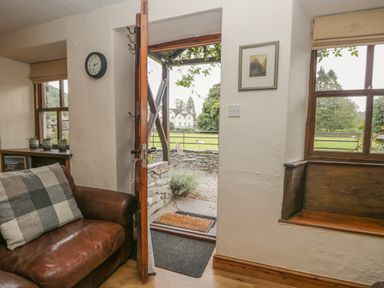 An open door leading outside with a brown leather sofa and a window with wooden bench seating at Tanner Croft Cottage in Grasmere
