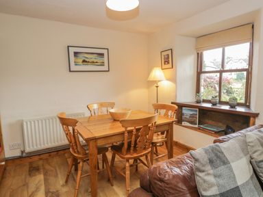 A dining area with a wooden table and four chairs next to a window with small plants and books at Tanner Croft Cottage in Grasmere