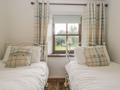 A bedroom with two single beds separated by a window with curtains and a bedside lamp at Tanner Croft Cottage in Grasmere