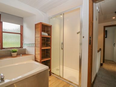 A bathroom with a bathtub a shower cubicle a wooden shelf with towels and a window with a blind at Tanner Croft Cottage in Grasmere