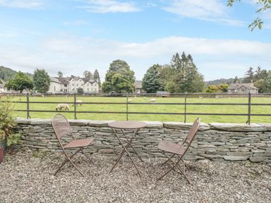 An outdoor seating area with two chairs and a round table on gravel by a stone wall overlooking a field with sheep at Tanner Croft Cottage in Grasmere