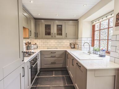 A kitchen with grey cabinets white countertops a gas stove an oven a sink and a window at Tanner Croft Cottage in Grasmere