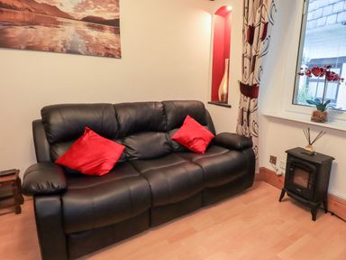 A black leather sofa with two red cushions next to a window and a small black stove at Granary Nook in Windermere