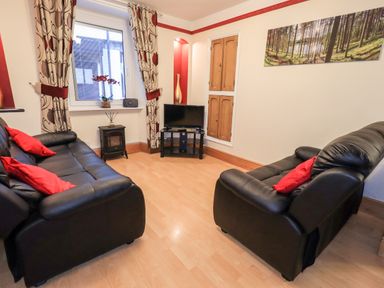A living room with black leather sofas and red cushions a small TV on a stand and curtains with a floral pattern at Granary Nook in Windermere