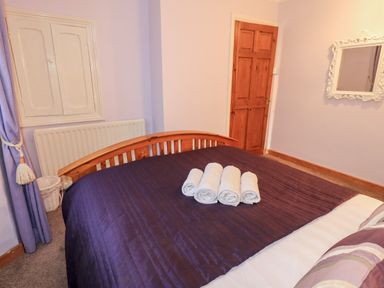 A bedroom with a wooden bed with dark bedding and rolled towels on the bed a wooden door a mirror on the wall and a window with curtains at Granary Nook in Windermere