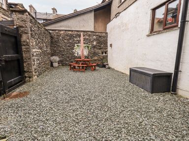 A courtyard with a gravel ground a wooden picnic table with an umbrella stone walls a plastic storage box and several potted plants at Granary Nook in Windermere