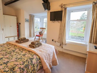 A bedroom with a bed with folded towels a rocking chair a wardrobe a wall-mounted TV and windows overlooking a garden at Greta Mill in Keswick
