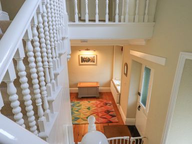 A staircase with white balustrades overlooking an entrance hallway with a colorful geometric rug a wooden chest a wall painting and a white door at Greta Mill in Keswick