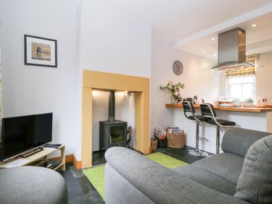 A living room with grey sofas a television a wood stove and a kitchen counter with two bar stools at Banks Place in Keswick