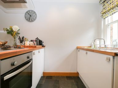 A kitchen with wooden countertops white cabinets a stove a clock on the wall and a window with patterned blinds at Banks Place in Keswick