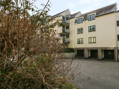 An exterior view of an apartment building with balconies and garages at ground level surrounded by leafless bushes at Tree Tops View in Bowness-On-Windermere