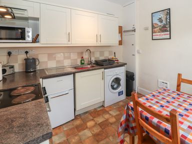 A kitchen with white cabinets, a washing machine, a small refrigerator, a sink, a microwave, a kettle, a table with a multicolored checkered tablecloth, and two wooden chairs at Pelham House in Keswick