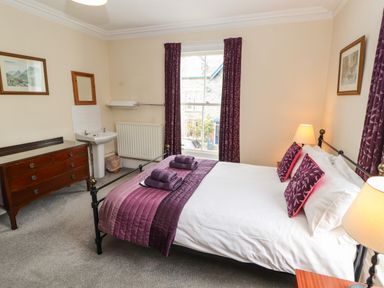A bedroom with a bed, bedside table with lamp, chest of drawers, sink, window with curtains and framed pictures at Pelham House in Keswick