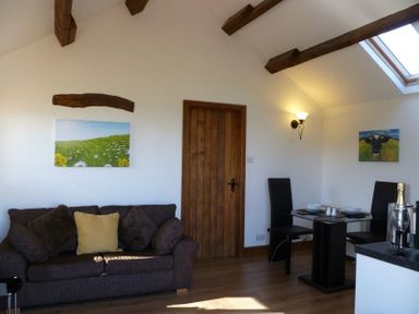 A sitting area with a dark sofa and cushions next to a wooden door and a dining table with two black chairs at Buttercup Cottage in Catterlen