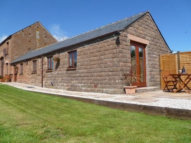 An exterior view of a stone cottage with brown framed windows a grass lawn and a small patio with wooden table and chairs at Buttercup Cottage in Catterlen