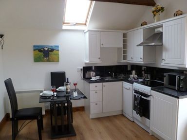 A kitchen with white cabinets black countertops a dining table set for two with wine glasses and plates at Buttercup Cottage in Catterlen