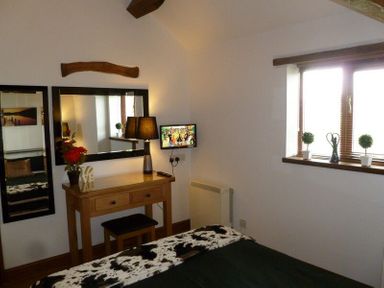A bedroom with a wooden dressing table stool lamp wall mirror small mounted TV and window with three potted plants at Buttercup Cottage in Catterlen