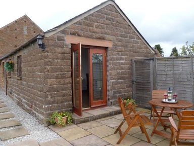 A stone exterior with wooden door opening to a patio with wooden table and chairs and a bottle of wine at Buttercup Cottage in Catterlen