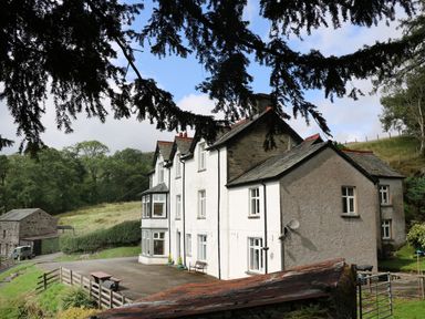 A white multi-story house with pitched roofs in a rural setting surrounded by trees and grassy hills at Low Skelgill in Troutbeck