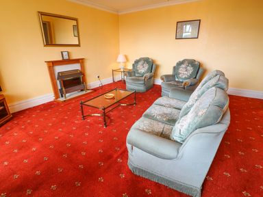 A living room with green upholstered sofa and chairs a glass coffee table a fireplace with a mirror above and a red patterned carpet at Low Skelgill in Troutbeck