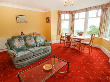 A living room with a green floral sofa a glass coffee table a wooden dining table with four chairs and large windows at Low Skelgill in Troutbeck