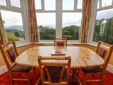 A dining area with a wooden table and four chairs by large windows with curtains at Low Skelgill in Troutbeck