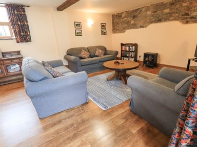 A living room with three sofas a round wooden table a bookshelf and a small stove at Tithe Cottage in Cockermouth