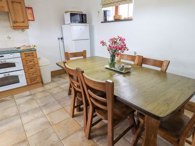 A kitchen with a wooden dining table and six chairs flowers in a vase on the table a refrigerator and microwave in the background at Tithe Cottage in Cockermouth